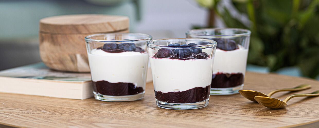 Trzy szklane pojemniki Viggo z jogurtem i warstwą ciemnej frużeliny jagodowej, udekorowane świeżymi jagodami na drewnianym stole